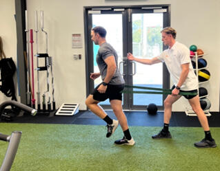 An exercise physiologist at Inertia Health Group assists a client with resistance-based exercises, focusing on lower-body strength, flexibility, and movement.